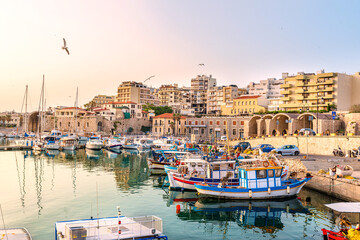 Fototapeta na wymiar View to Heraklion in the early morning during sun rise, the old port with traditional fishing boats and town with its antient buildings at the background. Selective focus