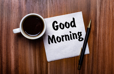 On a wooden table next to a white cup of coffee and a pen is a white paper napkin with the words GOOD MORNING.