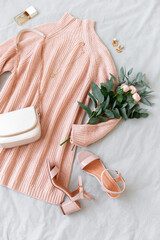 Pink knitted sweater dress, small bag with chain strap, heeled sandals, bouquet of roses and eucalyptus on gray background. Women's stylish spring outfit. Trendy casual clothes. Flat lay, top view.