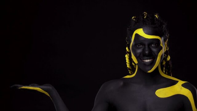 Face art. Girl pointing on copy space with fingers. Woman with black and yellow body paint. Young african girl with colorful bodypaint. An amazing afro american model with makeup.
