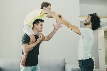 Two joyful dads and son having fun at home, boy riding on mans neck. Side view. Family and...