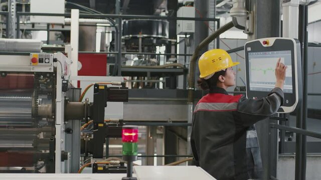 Slow-motion medium side-view shot of female mixed race engineer in uniform and safety helmet setting parameters for machine equipment on touchscreen at industrial plant