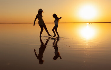 Two happy lovely sisters are walking along the mirror salt lake enjoying the evening fiery sunset