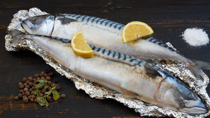 Two atlantic mackerel fishes ready for cooking decorated with spices - basil leaves and pepper, salt and lemon slices on the black wooden table 