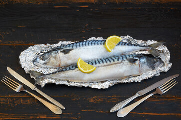 Two atlantic mackerel fishes ready for cooking decorated with spices, salt and lemon slices on the black wooden table close up