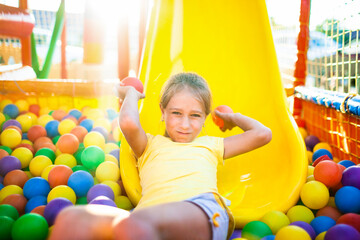 A cute cheerful girl lies on the playground with soft and bright equipment and throws colorful balloons into the camera, enjoying the warm summer sun