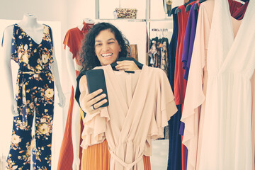 Joyful woman shopping in clothes store and consulting friend on cellphone, showing dress on hanger...