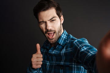 Joyful handsome guy winking and showing thumb up while taking selfie