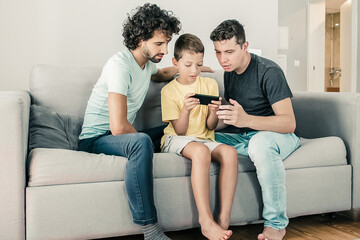 Serious boy playing online game on cellphone, his two dads sitting near him and looking at screen. Family at home and communication concept