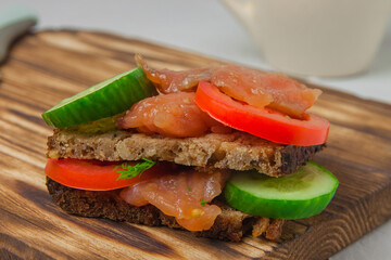 Red fish sandwich with wholegrain bread, cucumber, herbs and tomatoes laying on a wooden board