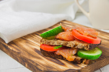 Red fish sandwich with wholegrain bread, cucumber, and tomatoes laying on a wooden board
