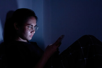 An Asian woman wearing glasses is using a smartphone with a bright screen in the dark.