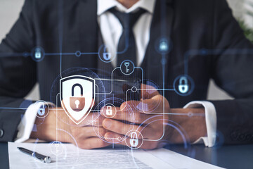 A businessman in formal wear reading the contract to prevent probability of risks in cyber security and checking details in the phone. Padlock Hologram icons over the working desk.