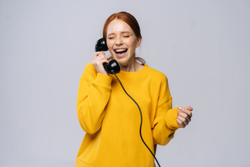 Laughing happy young woman in stylish yellow sweater talking on retro phone against isolated white background. Pretty redhead lady model emotionally showing facial expressions.