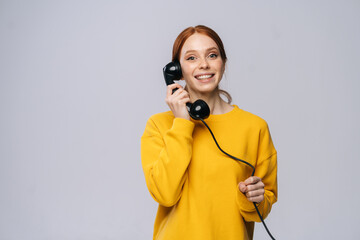 Cheerful young woman in stylish yellow sweater talking on retro phone and looking at camera against isolated white background. Pretty redhead lady model emotionally showing facial expressions.