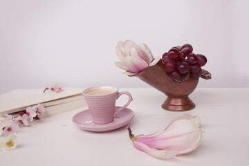 cup of cappuccino on a white table, blooming magnolia buds, a book in a white cover on a delicate pink background, a blank for the designer, an invitation form, a spring mood concept