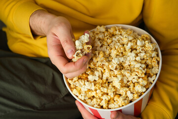 Popcorn paper bucket in the hands of a men preparing to watch a movie. Showtime. Eating delicious unhealthy sweet snacks. Going to cinema for a new film. Rest and entertainment. Popcorn closeup