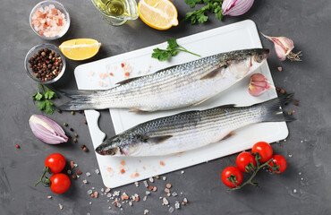 Raw fish mullet with ingredients and seasonings on white plastic board on dark background. View from above