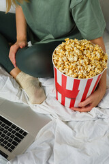 Popcorn bucket in the hands of a young girl preparing to watch a movie in bed on the laptop. Showtime. Eating delicious sweet snacks. Watching new film in home. Rest and entertainment in bedroom