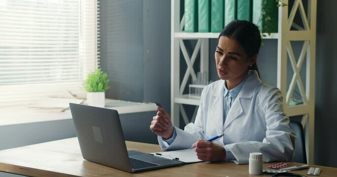 Female doctor consulting patient online through video call on laptop, listening to person's complaints and writing notes. Telemedicine concept.