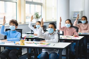 Schoolchildren raising hands at classroom, wearing medical masks