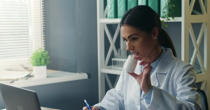 Smiling friendly female doctor using laptop for consulting patient online, holding pills bottle and explaining prescribed treatment. Close-up view.