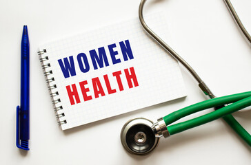 WOMEN HEALTH is written in a notebook on a white table next to pen and a stethoscope.