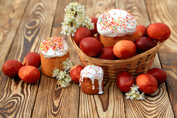 Easter eggs and Easter cupcake in a wicker basket on a wooden background with blooming sprigs of wild plum. 
Greeting card