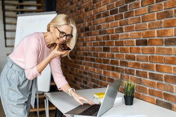 Confident middle aged businesswoman in glasses using voice recognition function on a smartphone. Modern female entrepreneur looking at the laptop, sending a vocal message via online messenger