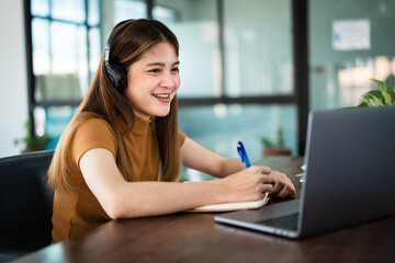 Young smile Asian girl student wears wireless headphones write on the notebook to study language online watch and listen to the lecturer, webinar via video call e-learning at home, distance education