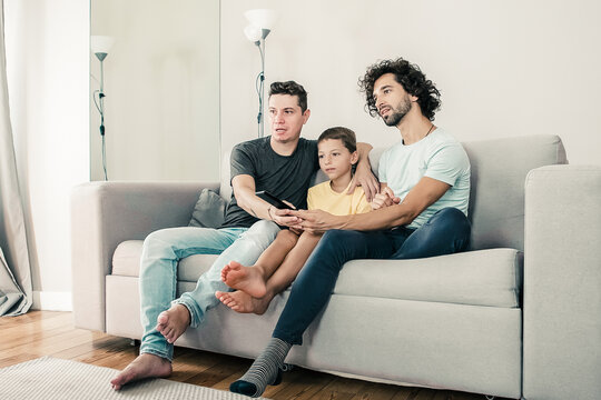Young handsome gay couple and their son watching TV show at home, sitting on couch in living room, hugging, using remote control, looking away. Family and home entertainment concept