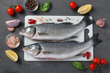 Raw seabass with ingredients and seasonings like basil, lemon, salt, pepper, cherry tomatoes and garlic on white plastic board on dark background. Top view