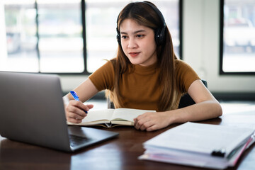 Obraz na płótnie Canvas Young Asian girl student wears wireless headphones concentrates write on the notebook to study language online watch and listen to the lecturer, webinar via video call e-learning at home