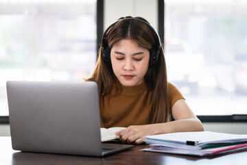 Young  Asian girl student wears wireless headphones concentrates write on the notebook to study language online watch and listen to the lecturer, webinar via video call e-learning at home