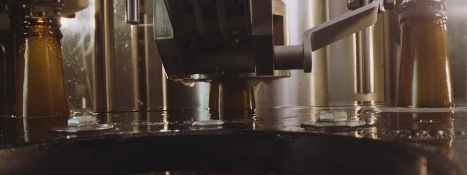 Close-up video of glass beer bottles being closed on a modern machine. Capping process of full beer bottles. Foam overflowing the beer bottles. Factory, brewery concept.