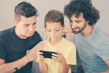 Focused cute boy using smartphone, his two dads sitting by him and looking at screen. Front view....
