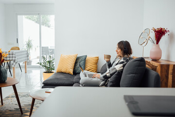 Concentrated and relaxed young woman sitting on comfortable couch, drinking a cup of coffee, working with a laptop at apartment. Freelancer or student working from home during Coronavirus pandemic.