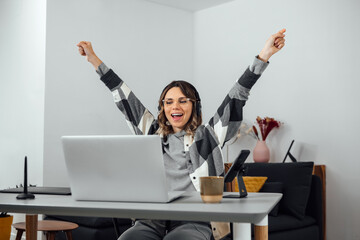 Excited woman, with arms opened, sitting at home office. Businesswoman freelancer Celebrating Success With Raised Hands. Overjoyed, euphoric, receiving good news at laptop, triumph, win lottery.