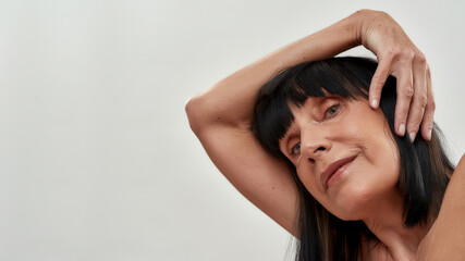 Fototapeta premium Mature beauty. Portrait of caucasian senior woman keeping one hand behind head, looking at camera while posing against grey background