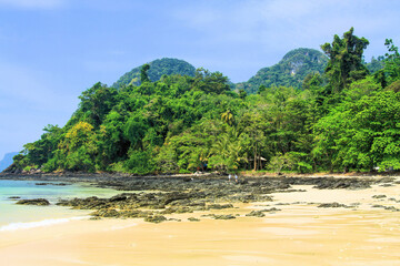 View over white sand and shallow water lagoon of tropical empty remote island beach with green hills jungle forest background - Koh Mook, Thailand