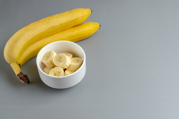 Two bananas and slices in a white plate on a gray background. Trending color Pantone of year 2021 Illuminating and Ultimate gray. Side view minimal still life with copy space.