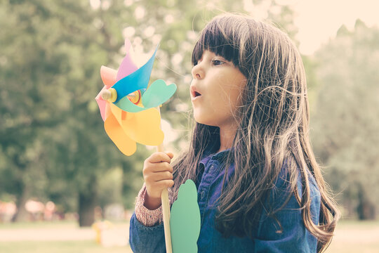 Adorable black haired girl playing in park, holding pinwheel and blowing on toy. Medium shot, side view. Children outdoor activity concept