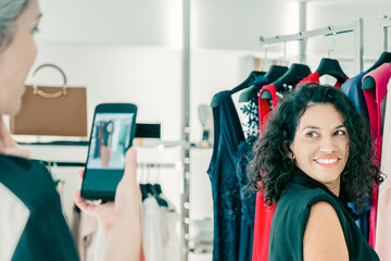 Happy female shoppers enjoying shopping in clothes store together, touching dress, posing and...