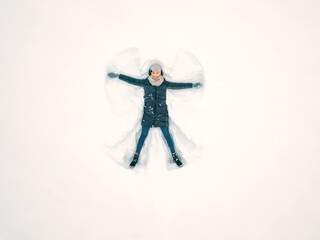 Young beautiful woman in a gray jacket, hat and gloves, blue jeans, boots makes a snow angel in the...