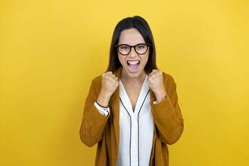 Young beautiful woman wearing a blazer over isolated yellow background very happy and excited making winner gesture with raised arms, smiling and screaming for success.