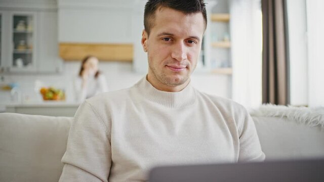 Close up concentrated self-isolated man working on laptop at home, his wife on background