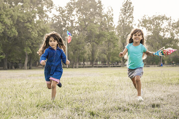 Joyful black haired little girls with pinwheels running on grass, having race in park. Front view, full length. Children outdoor activity concept