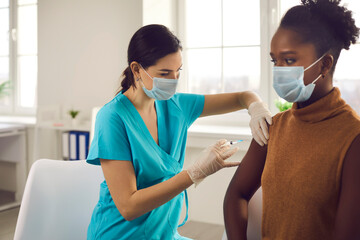 Professional nurse or doctor in medical face mask and gloves giving injection to female patient. Young African-American woman getting flu or modern Covid 19 vaccine shot during vaccination campaign - obrazy, fototapety, plakaty