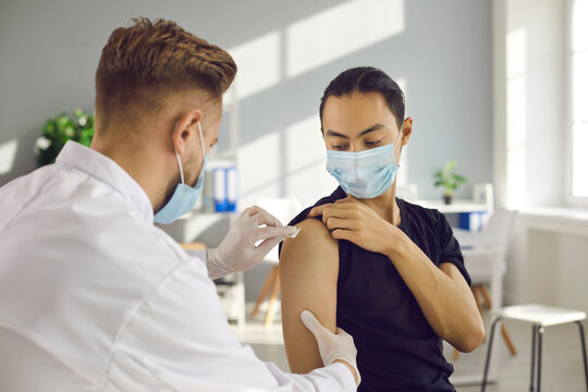 Young man in medical face mask getting seasonal flu shot during infection outbreak. Male doctor cleans skin on patient's arm before injecting modern Covid-19 vaccine during immunization campaign