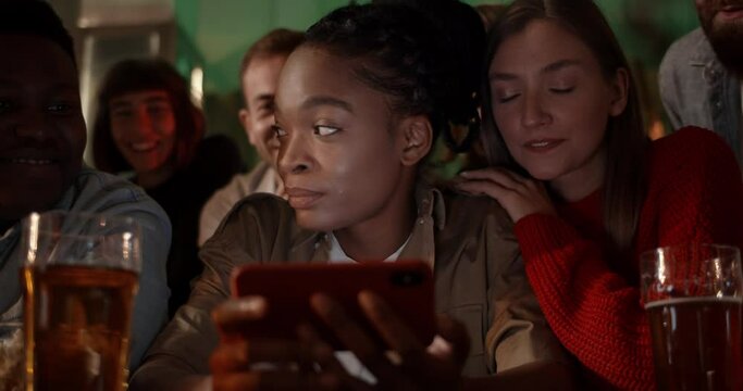 Crop view of afro american woman holding phone and smiling while sitting with friends in pub and looking at screen. Group of multiethnic people watching sport at smartphone